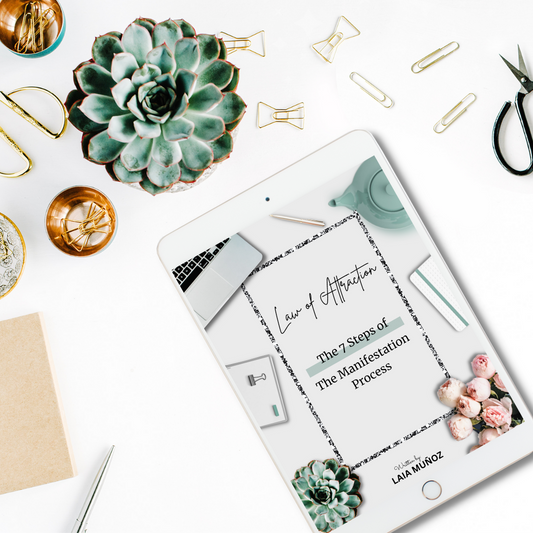 A PDF guide titled 'LAW OF ATTRACTION. The 7 Steps of the Manifestation Process' displayed on a tablet, with stationery items like pens, paper clips, and a succulent plant in the background and you can buy it at laiamunozauthor.com
